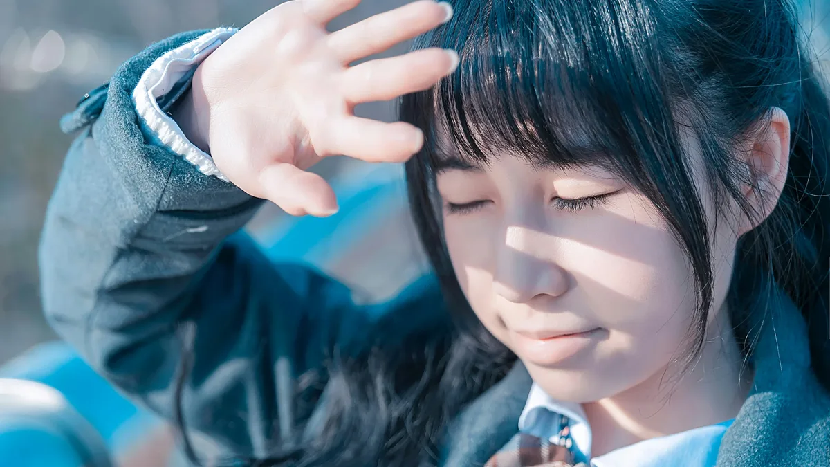 A young girl with long dark hair, wearing a school uniform, with her hand raised to her forehead.
